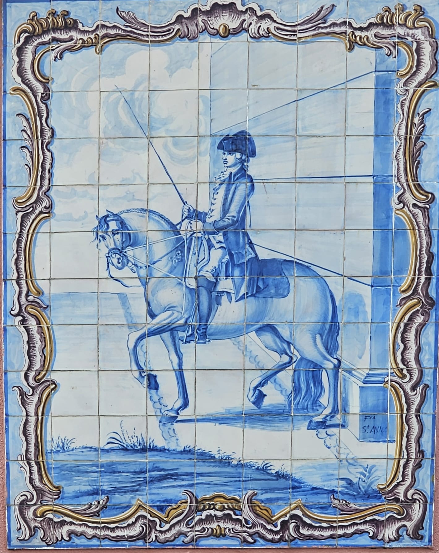 Lisbon tile. A detailed blue and white tile mural depicts a mounted rider in historical attire, holding a long whip. The horse is elegantly poised, set against a scenic backdrop with clouds. The artwork is framed by an ornate, gold-accented border.
