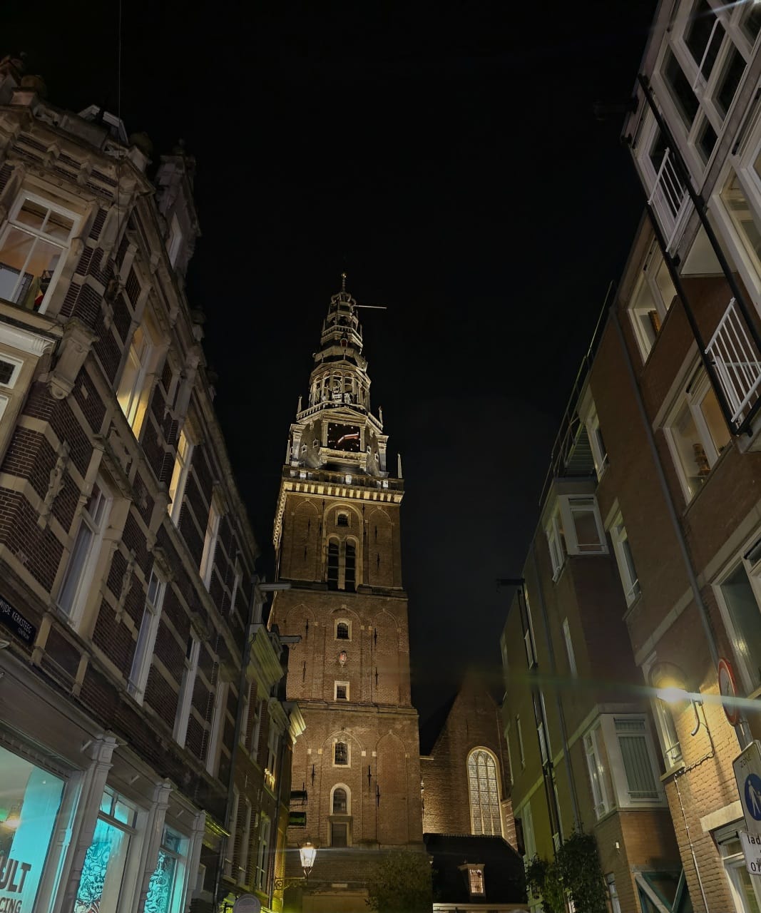 The photograph, taken from a narrow street looking up, captures the illuminated brick tower and spire of the Oude Kerk, Amsterdam's oldest building and parish church.