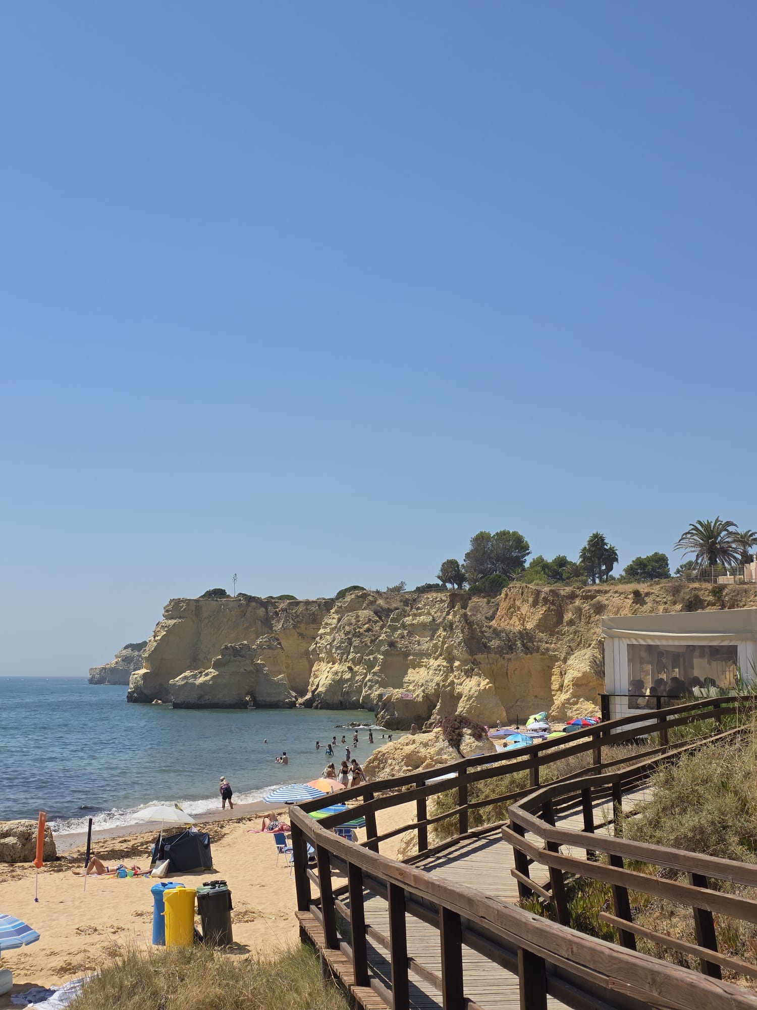 A sunny beach scene at Praia do Vale do Olival in Portugal, showing a wooden boardwalk leading down to a sandy beach backed by golden cliffs and calm turquoise water. People are relaxing on the sand and swimming in the sea