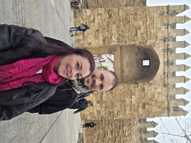 A smiling couple stands in front of a large, very old wall and gate in Cordoba, Spain
