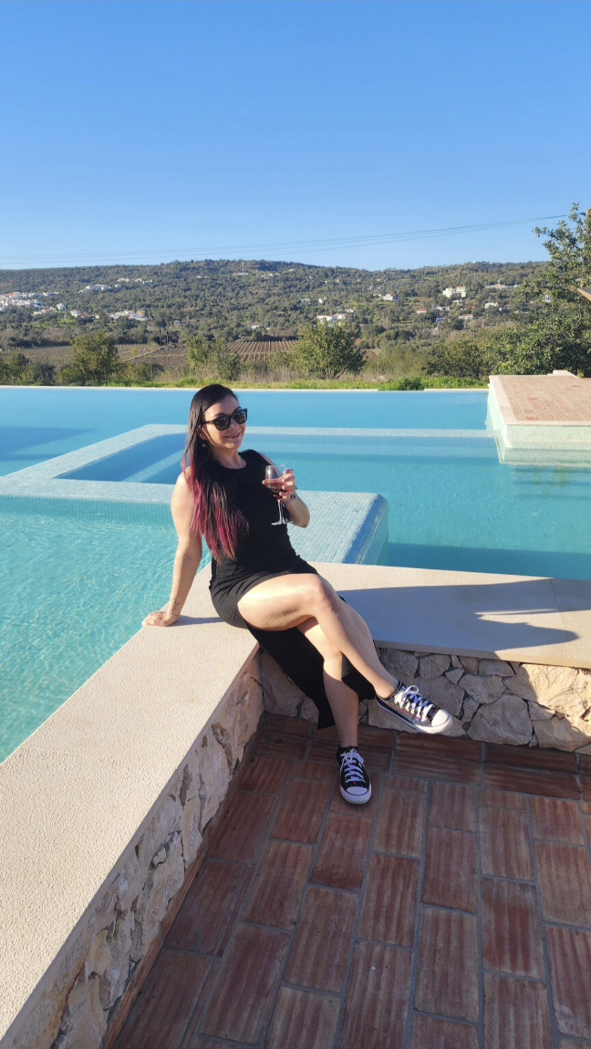 A woman site by an infinity pool, holding a glass of wine. There are mountains behind her