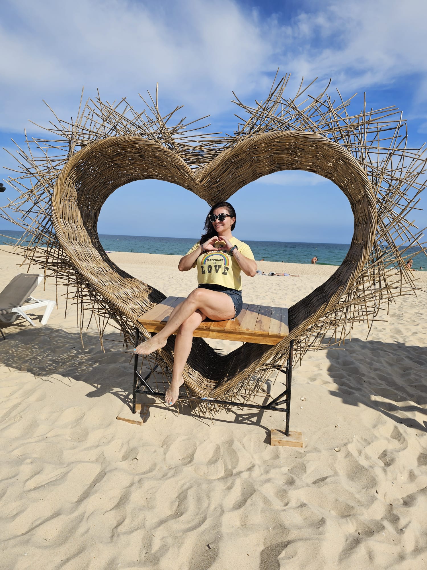 A woman sits on a wooden bench inside a large, heart-shaped sculpture made of woven sticks, located on a sandy beach. The ocean is visible in the background under a partly cloudy sky
