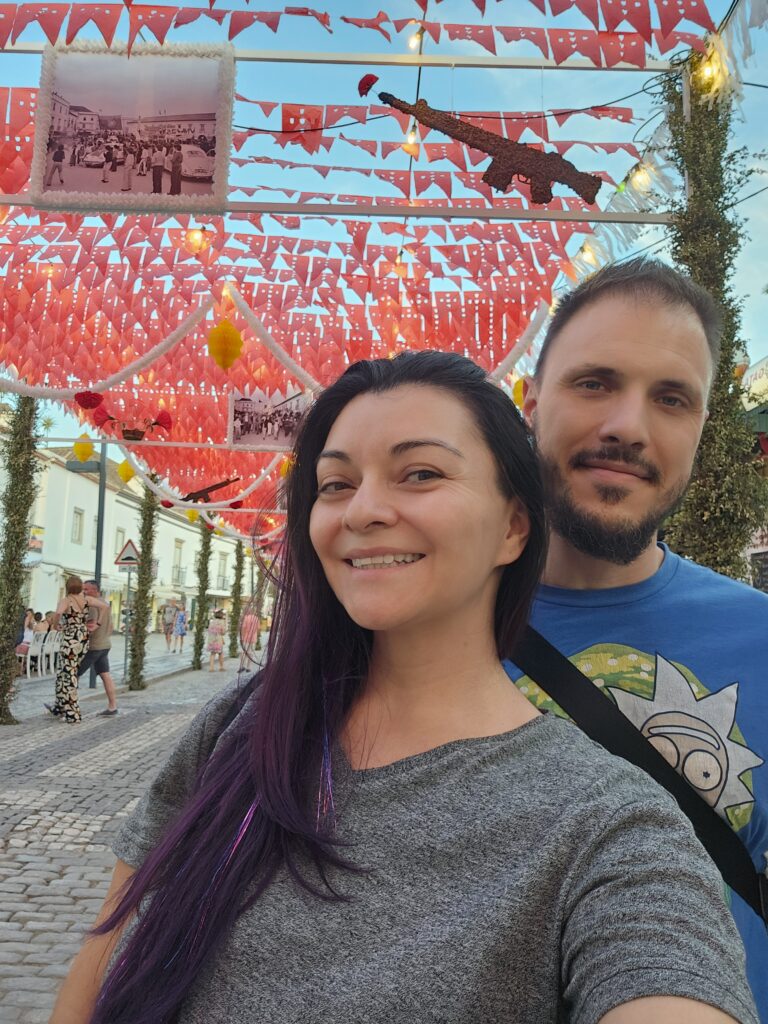A couple taking a selfie in front of elaborate red paper flag decorations strung across a cobblestone street in Tavira, Portugal during Santos Populares