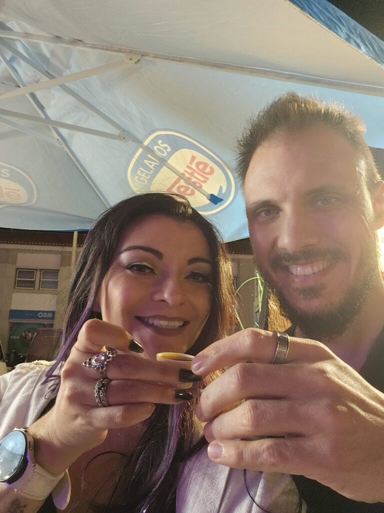 A couple smiling and holding up small shot glasses at a Santos Populares street festival in Portugal, with a gelato vendor tent visible behind them