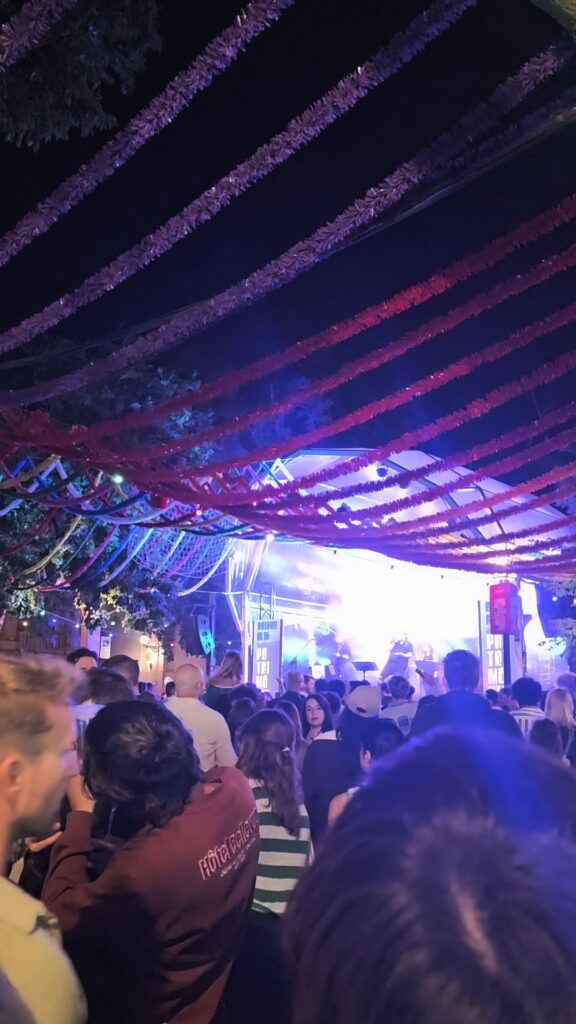 A large crowd gathered under colorful streamers watching a live band perform on a lit stage at a Santos Populares street party in Portugal at night