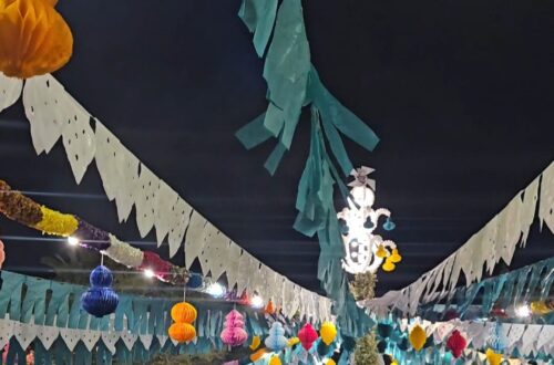 A crowd gathered at night beneath an elaborate canopy of colorful paper decorations, honeycomb lanterns, and bunting flags at a Santos Populares street party in Tavira, Portugal