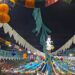 A crowd gathered at night beneath an elaborate canopy of colorful paper decorations, honeycomb lanterns, and bunting flags at a Santos Populares street party in Tavira, Portugal